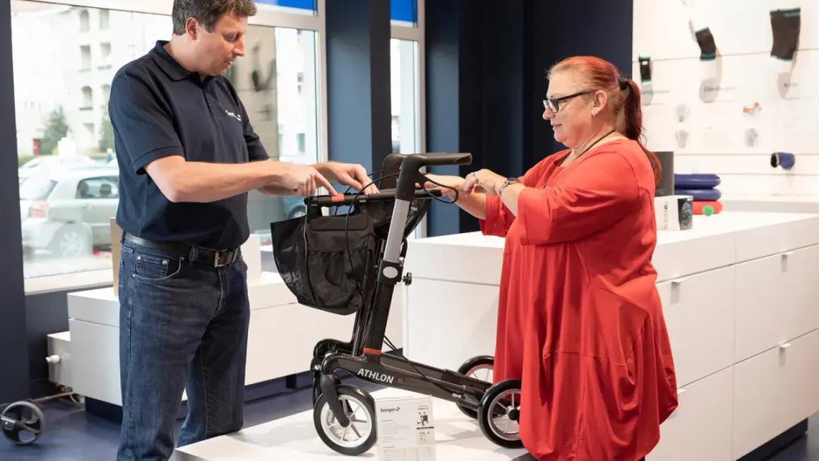 Mann erklärt einer Frau einen Rollator. Eine medizinische Hilfsmittel Liste ist im Hintergrund zu sehen.