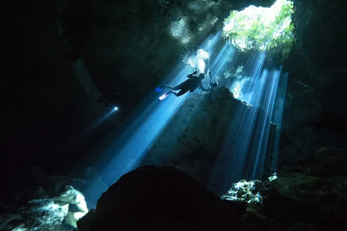 Buceadores exploran la asombrosa profundidad de un cenote, con rayos de luz solar atravesando la oscuridad.