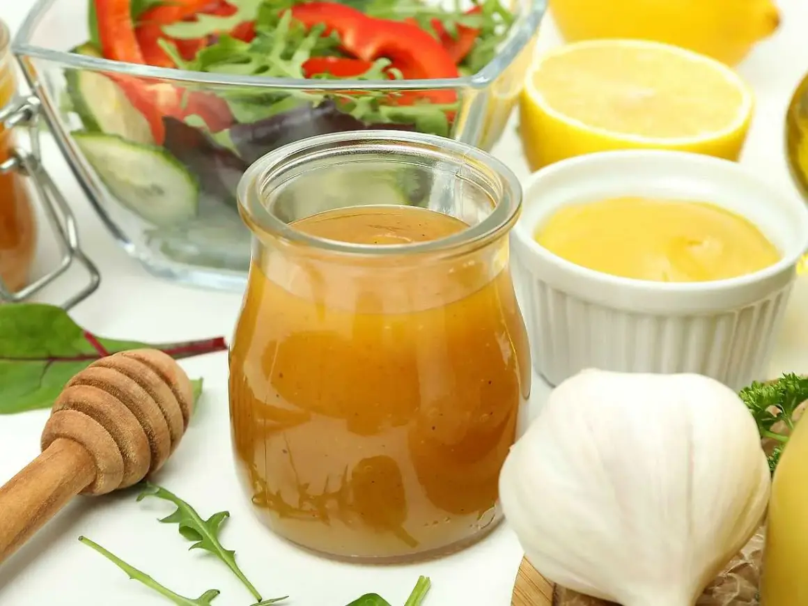 En burk med gyllene salladsdressing, bredvid en skål med sallad, citron, vitlök och honung. Ett perfekt salladsdressing recept!