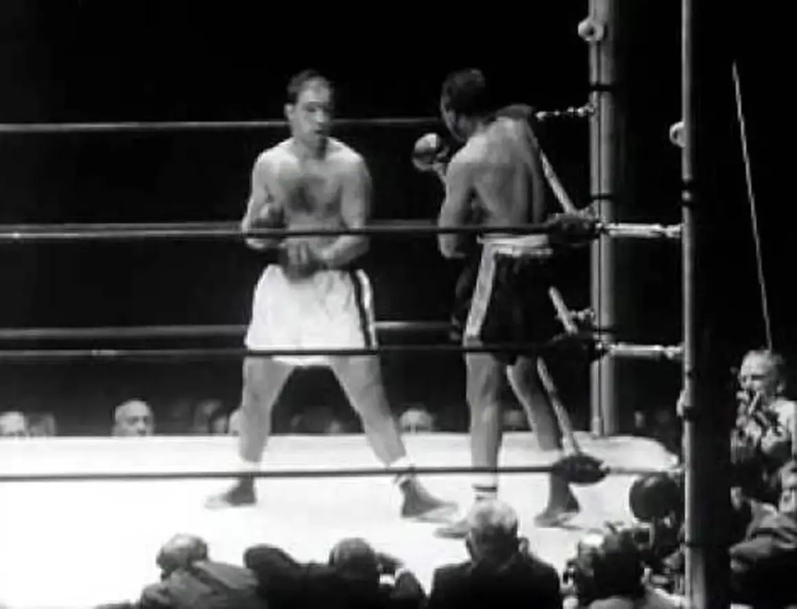Two boxers in a ring. One boxer, possibly Rocky Marciano, is in the foreground, his reach evident as he prepares to strike.