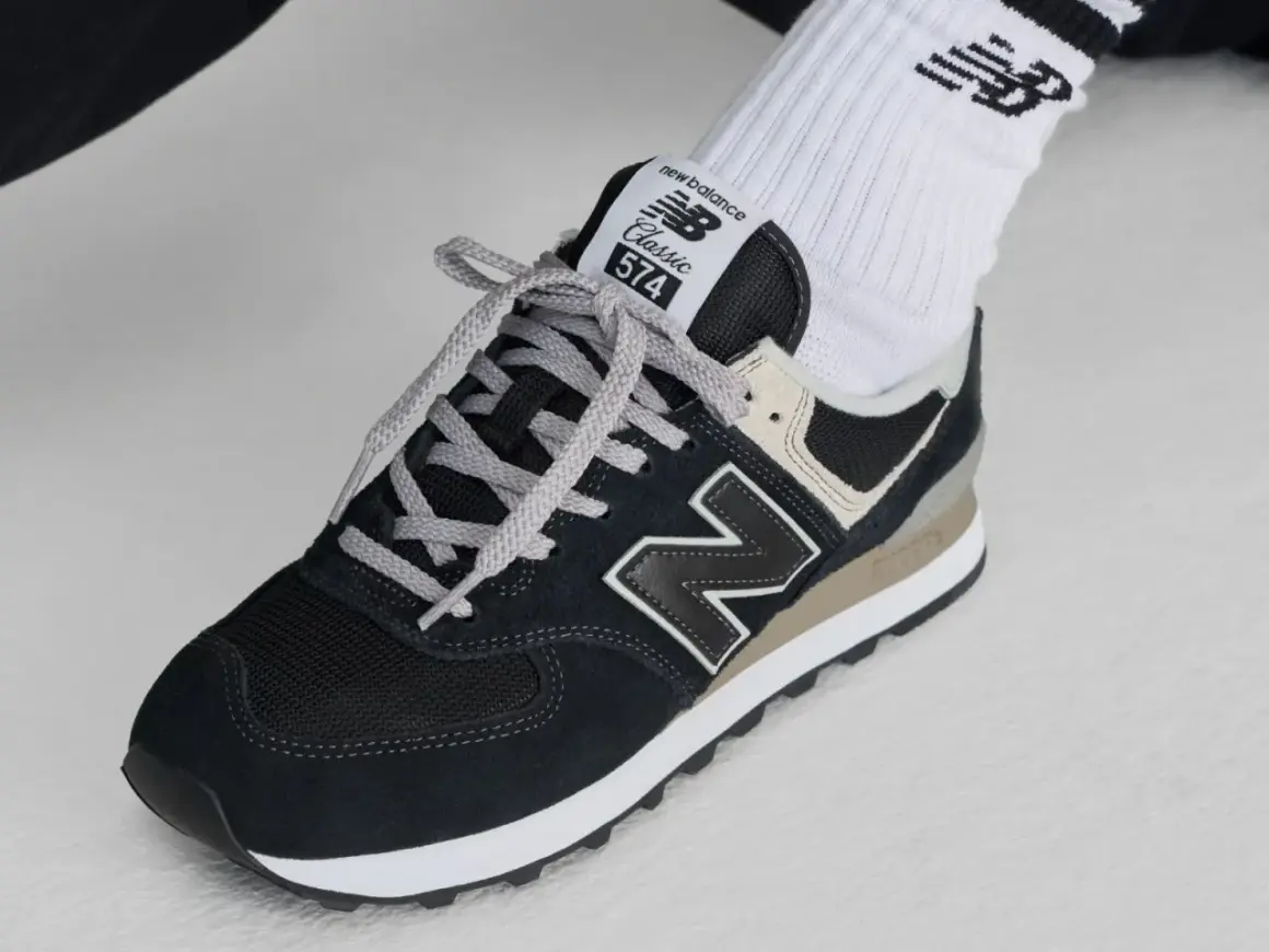 Klasyczne czarne buty sportowe new balance wl 574 z szarymi sznurówkami i beżowymi akcentami, noszone z białymi skarpetkami z logo.