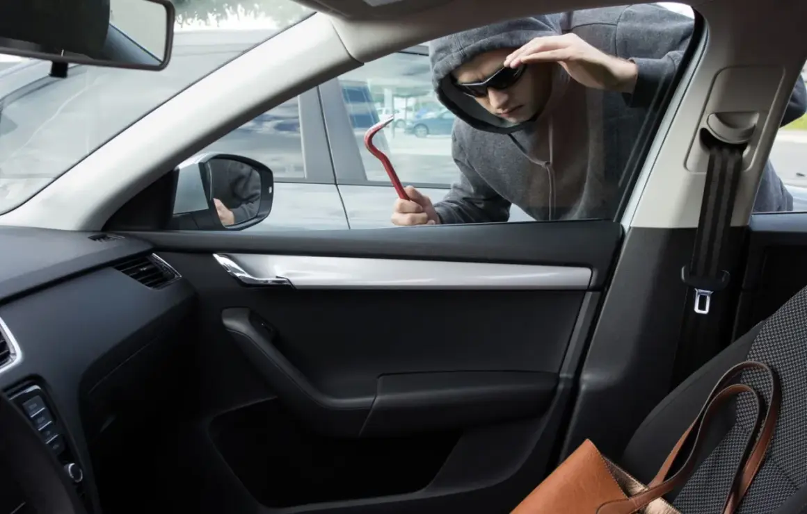 Un homme en capuche et lunettes de soleil tente un vol à la roulotte, outil rouge à la main, regardant à l'intérieur d'une voiture.