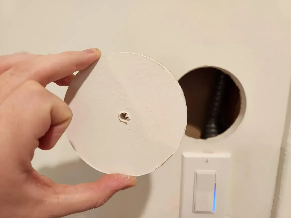Un disco di cartongesso viene tenuto in mano vicino a un buco nel muro, con un interruttore della luce visibile sotto.
