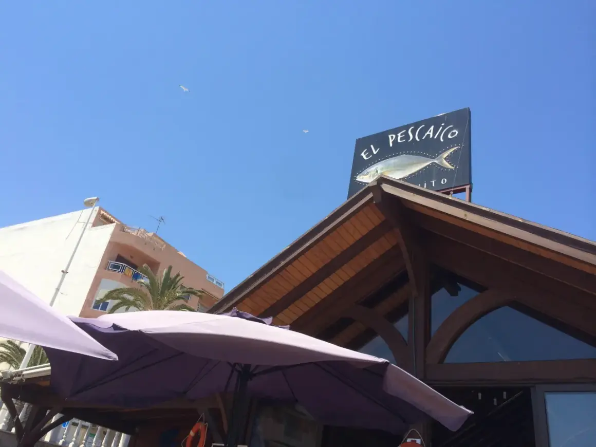 Un cartel de "El Pescaito" con un dibujo de pez, bajo un cielo azul con gaviotas.