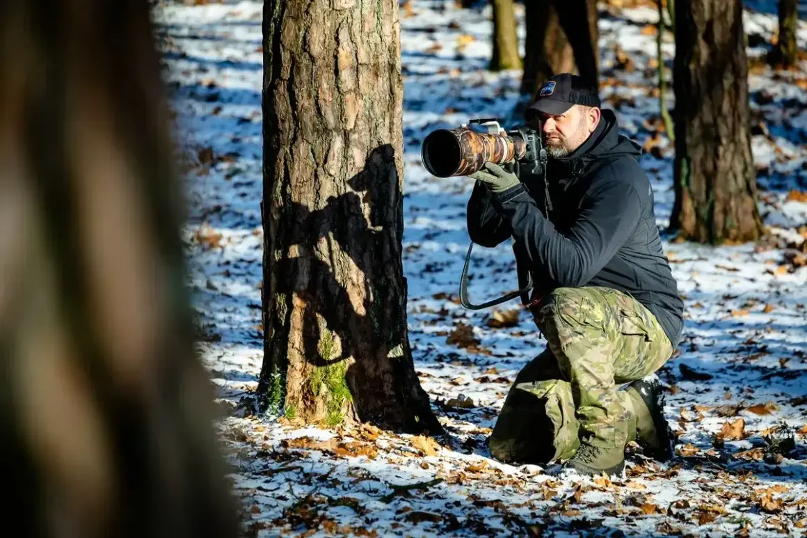 Fotografia przyrody: mężczyzna w kamuflażu kuca przy drzewie, celując profesjonalnym aparatem.