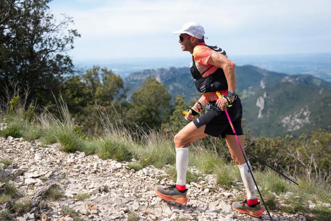 Uomo con bastoncini da trail running cos'è, corre su sentiero roccioso con vista panoramica su montagne e cielo azzurro.