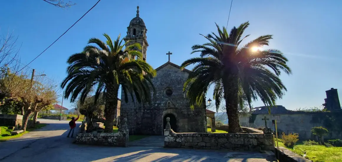 Iglesia de piedra con palmeras y sol brillante. Un lugar tranquilo que ver en Rianxo, ideal para pasear.