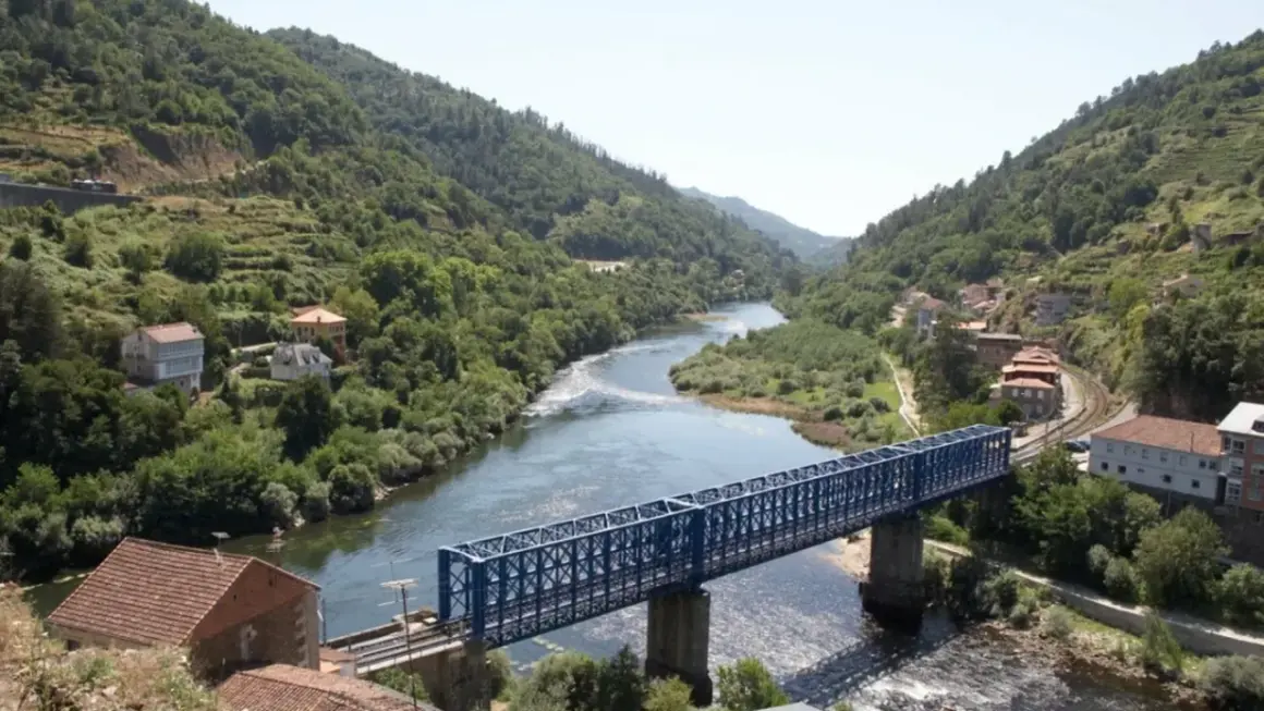 Un puente azul cruza un río serpenteante rodeado de colinas verdes y casas. El paisaje evoca la tranquilidad de los peares en un día soleado.