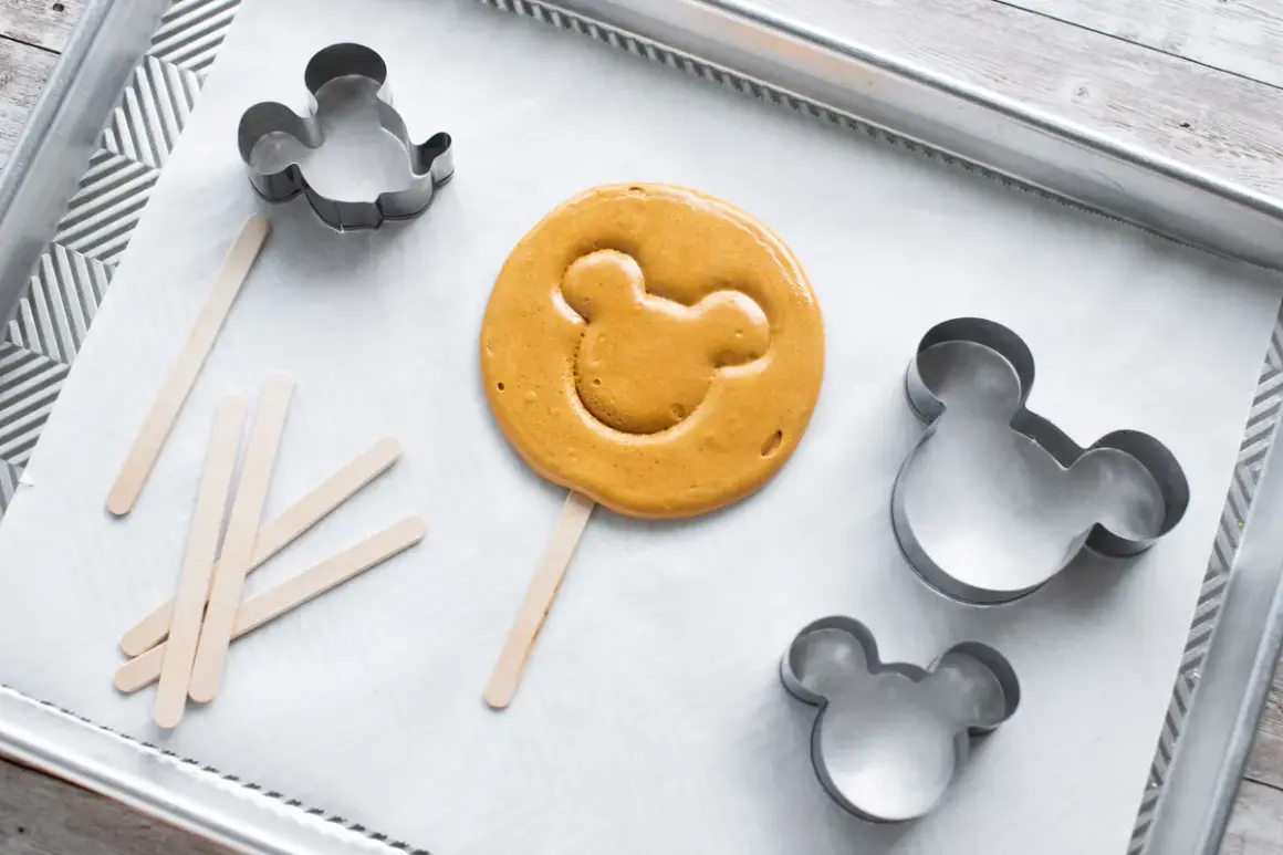 En färdiggjord dalgona cookie med Musse Pigg-form ligger på en bakplåt med flera Mickey Mouse-formar och träpinnar.