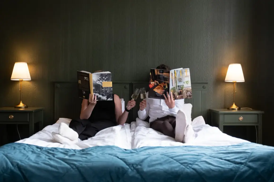 Två personer läser tidningar och skålar med champagne i en säng på Hotell Hertig Karl Filipstad.
