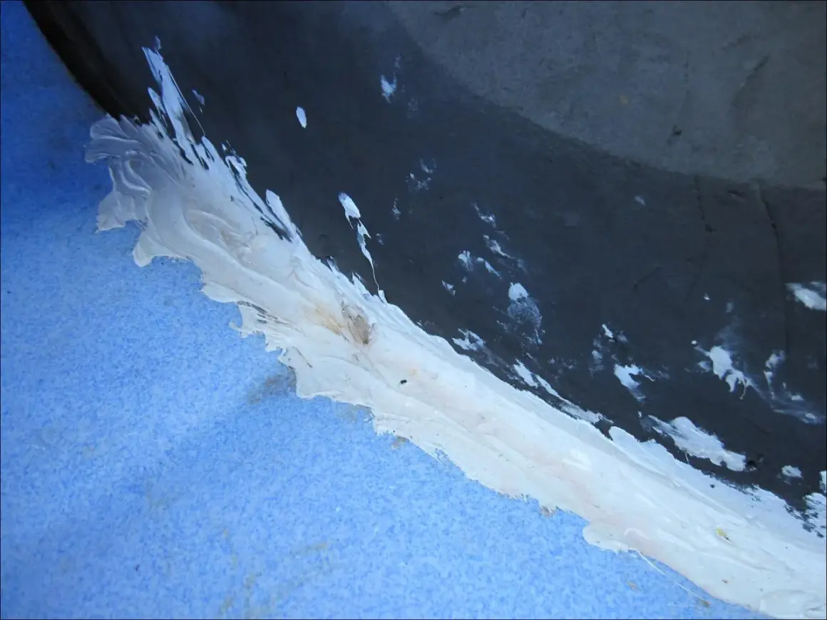 Application d'un mastic blanc pour coller du polyéthylène sur une surface bleue texturée, avec des éclaboussures sur le fond sombre.