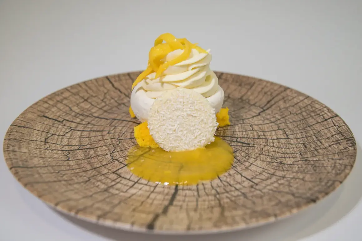 Deliciosa pavlova de mango con merengue, crema batida, coco rallado y salsa de mango sobre un plato rústico.