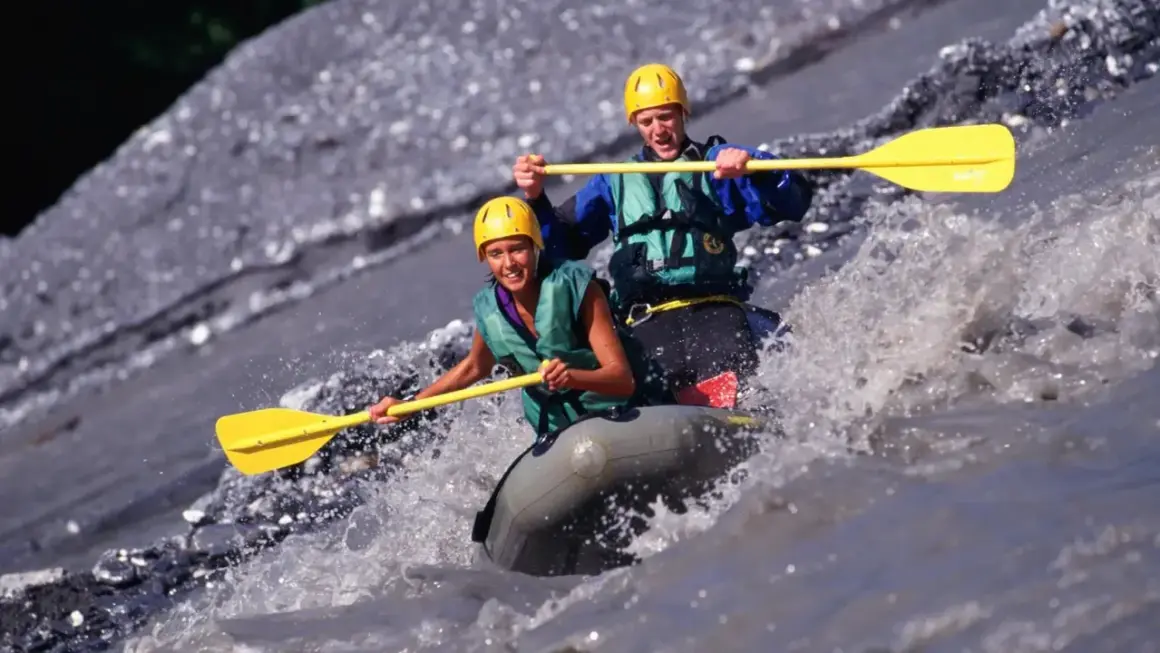 Dwóch uśmiechniętych ludzi w kaskach i kamizelkach płynie pontonem po rwącej rzece. To właśnie rafting – ekscytująca przygoda!