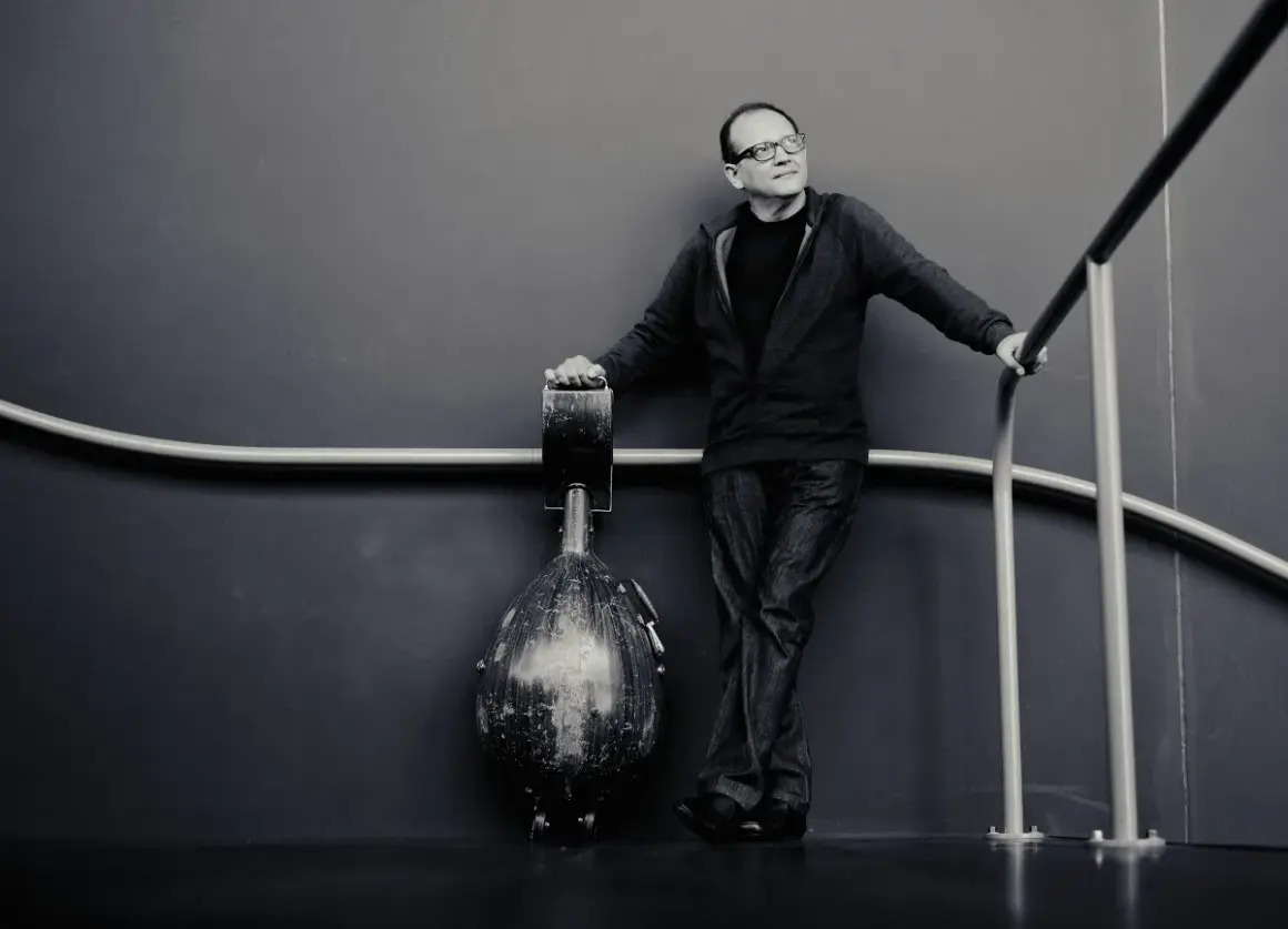 Anouar Brahem, w okularach, opiera się o poręcz, obok niego stoi instrument.
