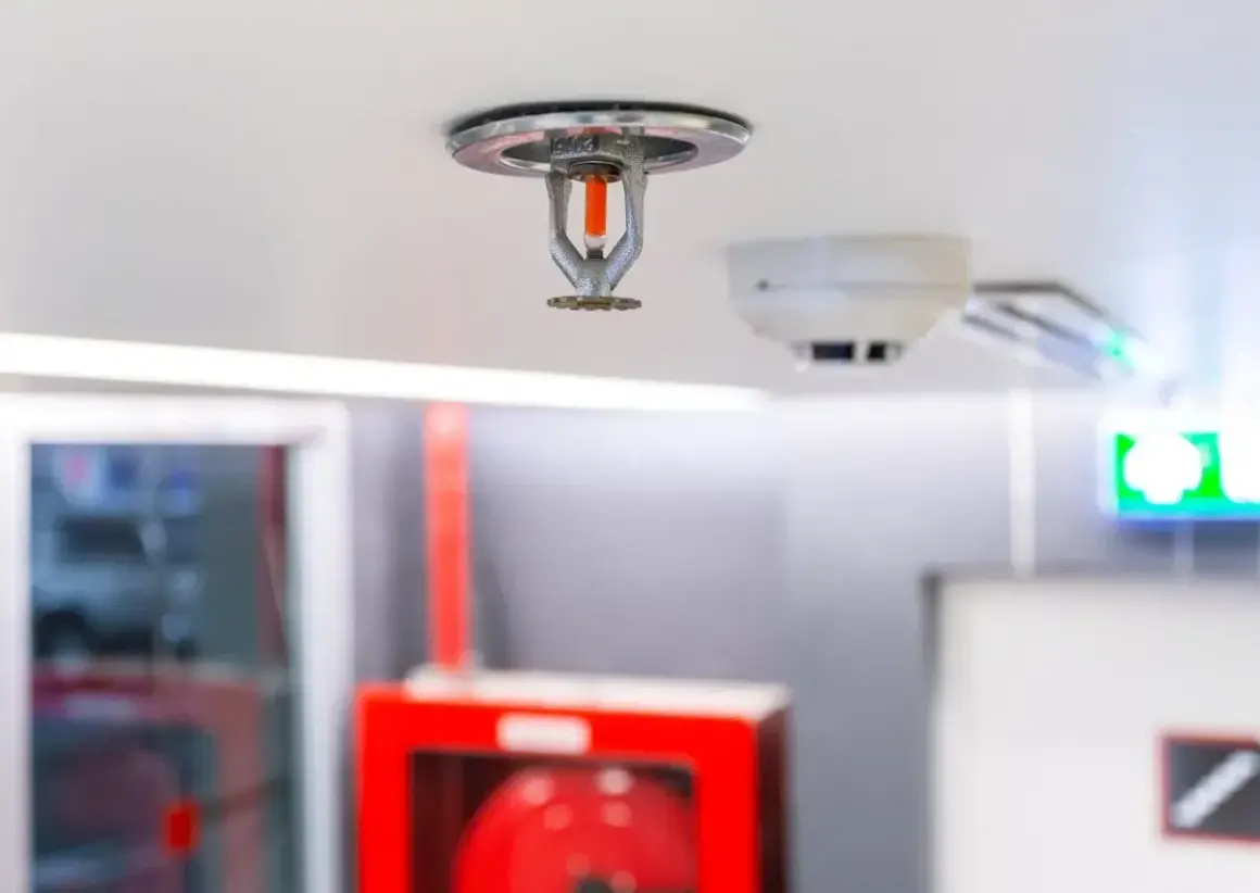 Eine Sprinkleranlage an der Decke, bereit für den Notfall. Daneben ein Rauchmelder und ein grünes Notausgangsschild.