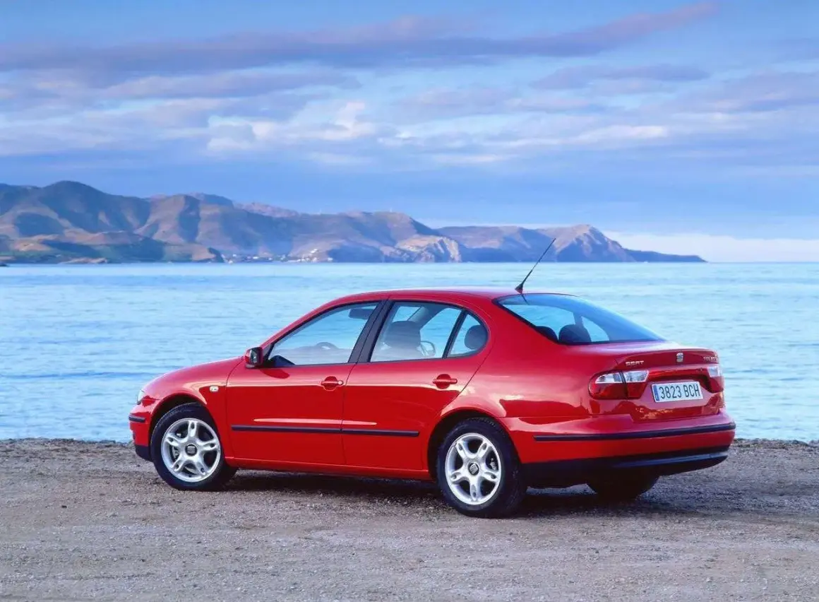 Czerwony Seat Toledo 2 na plaży, z widokiem na morze i góry w tle. Idealny na wakacyjną podróż.