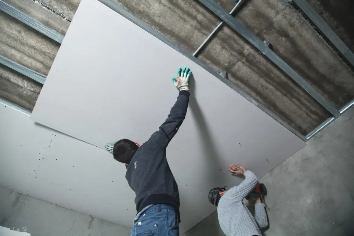 Deux ouvriers s'affairent à habiller un plafond abîmé avec des plaques de plâtre, fixées sur une structure métallique.
