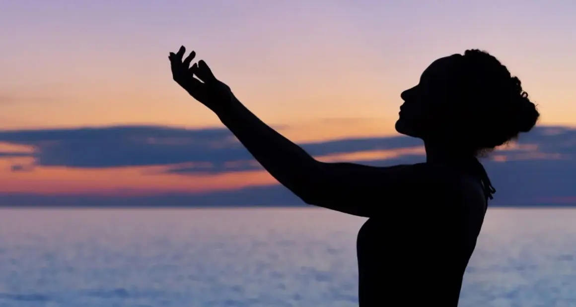 Silhouette einer Frau mit erhobenen Armen vor einem farbenfrohen Himmel und Meer. Sie scheint einen **chant deutsch** zu singen.