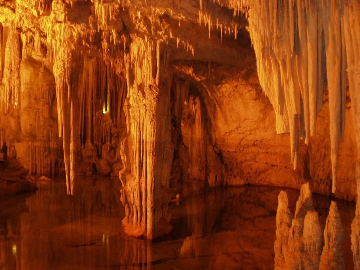 Interior de la cueva Rei Cintolo, con estalactitas y estalagmitas iluminadas en tonos cálidos, reflejándose en el agua subterránea.
