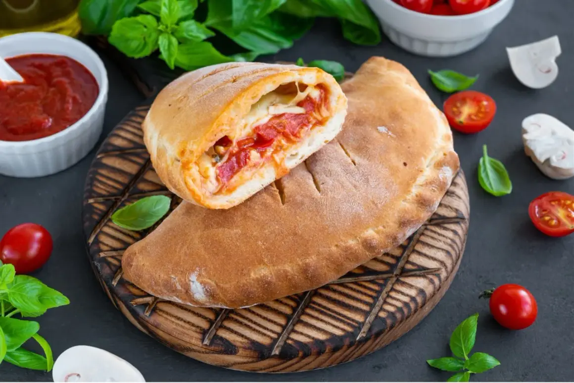 Złocisty calzone, przekrojony na pół, odsłaniający gorące nadzienie z pomidorów i sera. Obok sos pomidorowy i świeża bazylia.