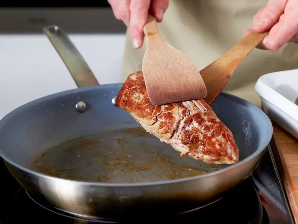 Ein saftiges Schweinefilet braten in der Pfanne. Mit zwei Holzspateln wird das Fleisch gewendet, um es von allen Seiten perfekt zu garen.
