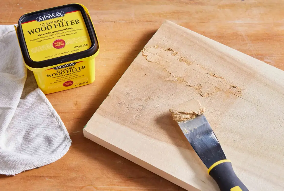 Un pot de mastic à bois Minwax est à côté d'une planche de bois où un couteau à mastic applique le produit pour reboucher bois.