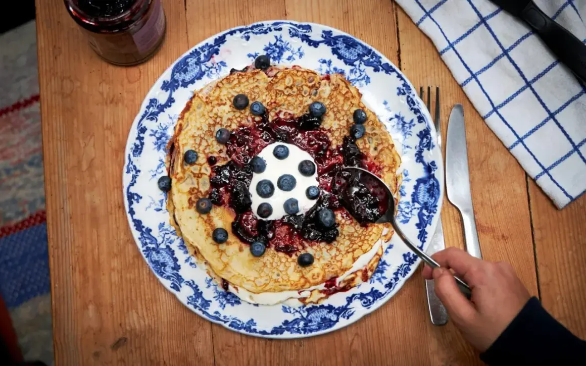 En hög med pannkakor toppas med grädde, blåbär och en generös klick drottningsylt. Perfekt för ett mysigt fika.