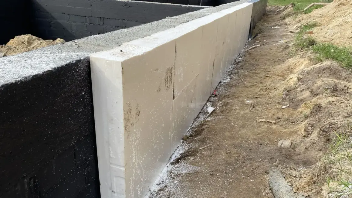Izolacja ze styropianu na czarnej ścianie fundamentowej. To dobry sposób, co położyć na fundament domu, by zapewnić mu ciepło.