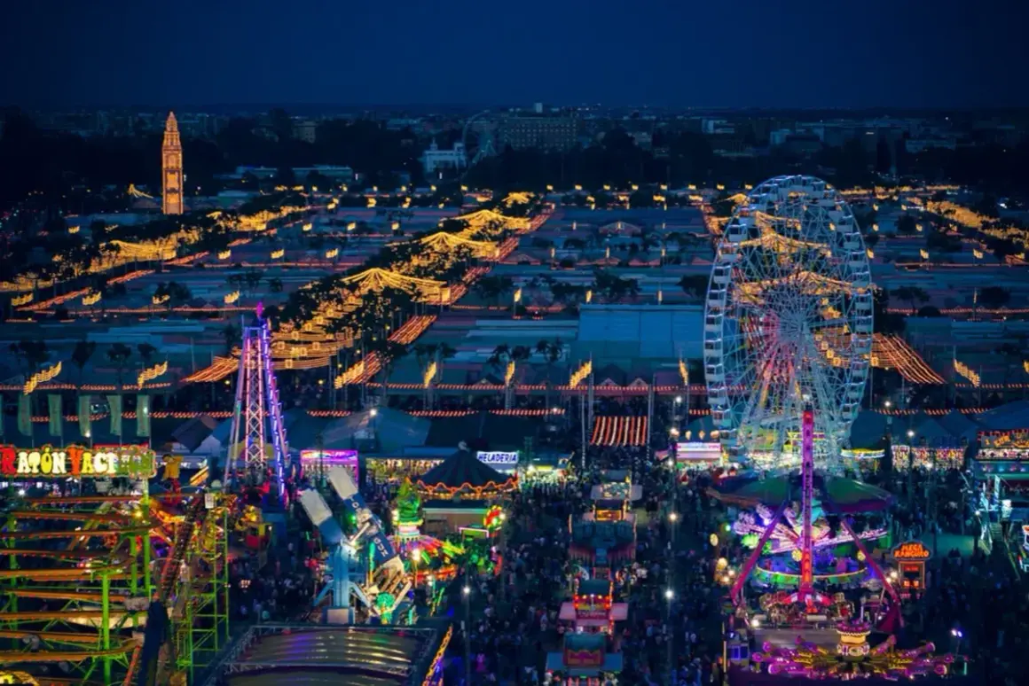 Nocne, rozświetlone atrakcjami wesołego miasteczka, gdzie trwa **feria de abril**. Widać diabelski młyn, kolejki górskie i tłumy ludzi.