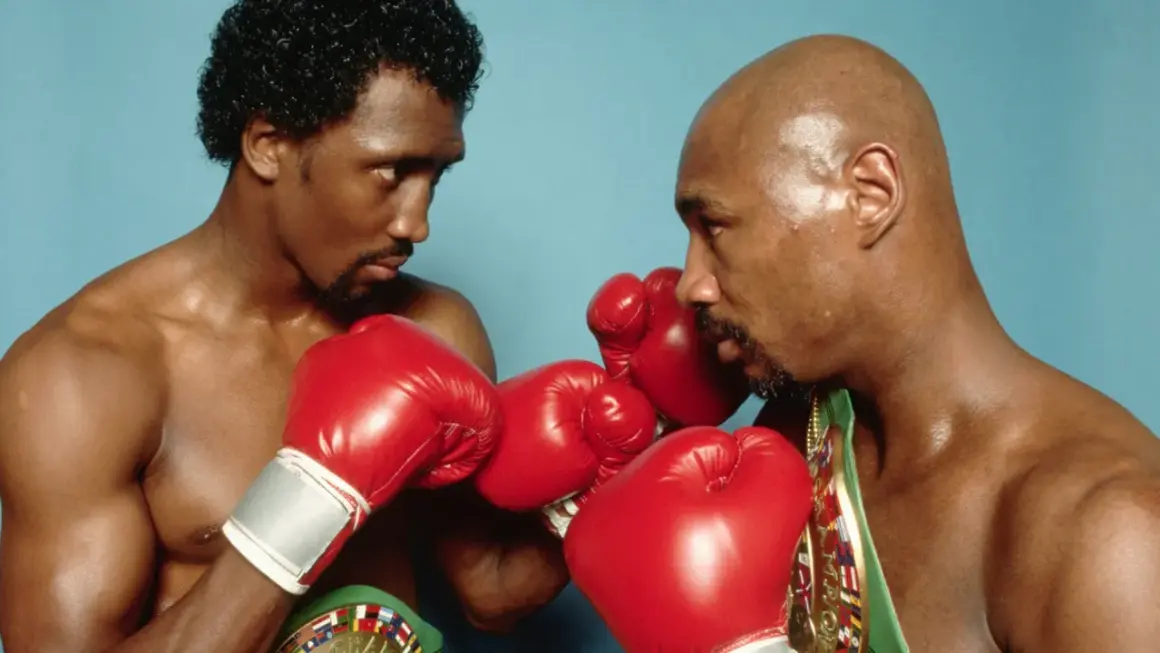 Two boxers, one with curly hair, the other bald, face off in a tense stare-down, red gloves raised, hinting at the legendary Marvin Hagler war.
