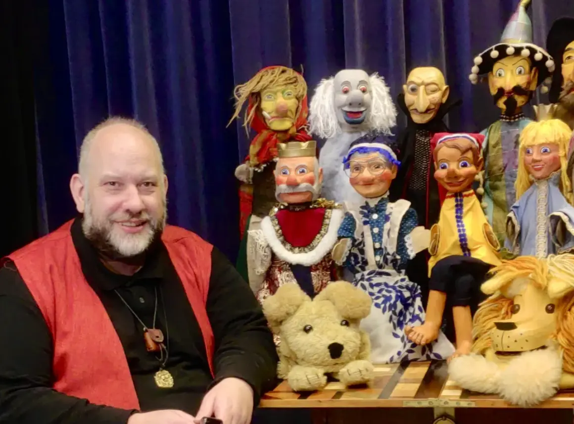 Ein Mann lächelt neben einer bunten Truppe von Handpuppen, die für den Puppenpavillon Bensberg bereitstehen.