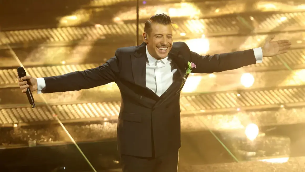 Un artista sorridente sul palco di Sanremo, con microfono in mano, pronto per i duetti.