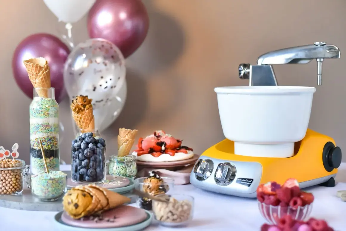 En gul och silverfärgad frozen yoghurt glassmaskin står redo för fest. Strössel, bär och våfflor väntar på att bli till godsaker.