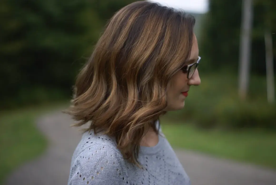 Une femme aux mèches brunes sur cheveux blancs, portant des lunettes, regarde au loin dans un décor verdoyant.