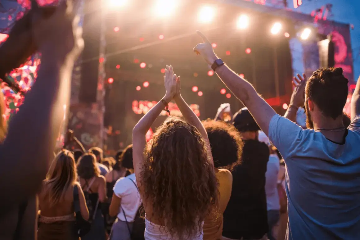 Folla entusiasta applaude e balla sotto le luci di un concerto. Per godersi al meglio l'evento, pensa a cosa portare ad un concerto: acqua, crema solare e un sorriso!