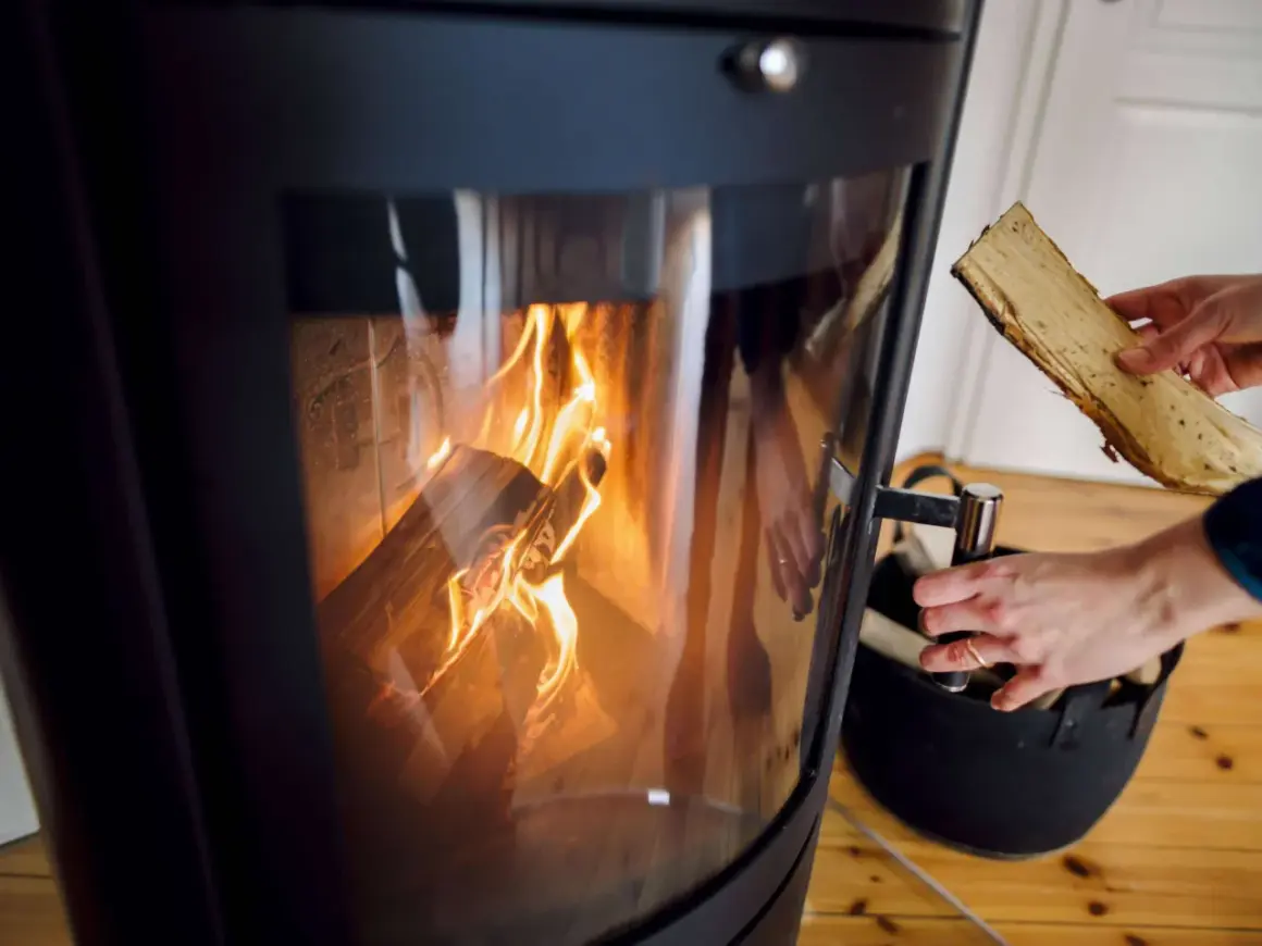 Ein Kaminofen mit brennendem Holz. Eine Hand hält ein Holzscheit, bereit zum Nachlegen. Bald muss der alte Kaminofen entsorgt werden.