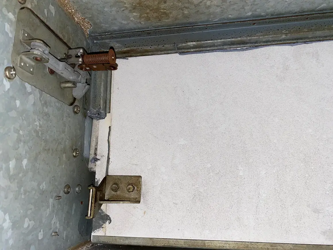 Detailaufnahme einer Brandschutzklappe mit sichtbaren mechanischen Teilen und einer weißen Platte. Möglicherweise Asbest-verdächtige Materialien.