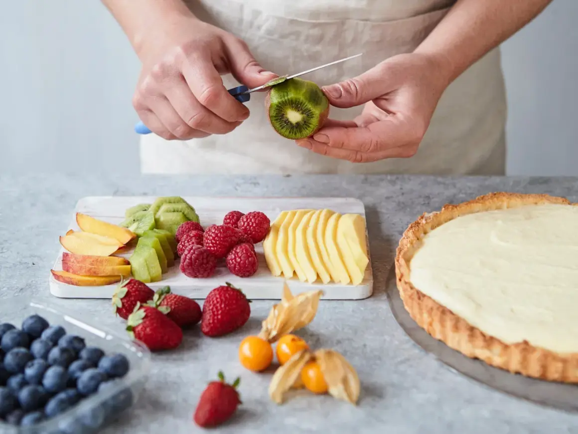 Hände schälen Kiwi für ein fruchtiges Tarte Rezept. Daneben liegen Beeren, Pfirsichspalten und Apfelscheiben.