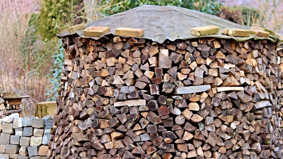 Ein großer Stapel Brennholz, der mit einer Plane und Ziegeln abgedeckt ist. Die Holzscheite sind ordentlich aufgeschichtet, bereit für den Winter. Die Baustahlmatte dient als stabile Unterlage.
