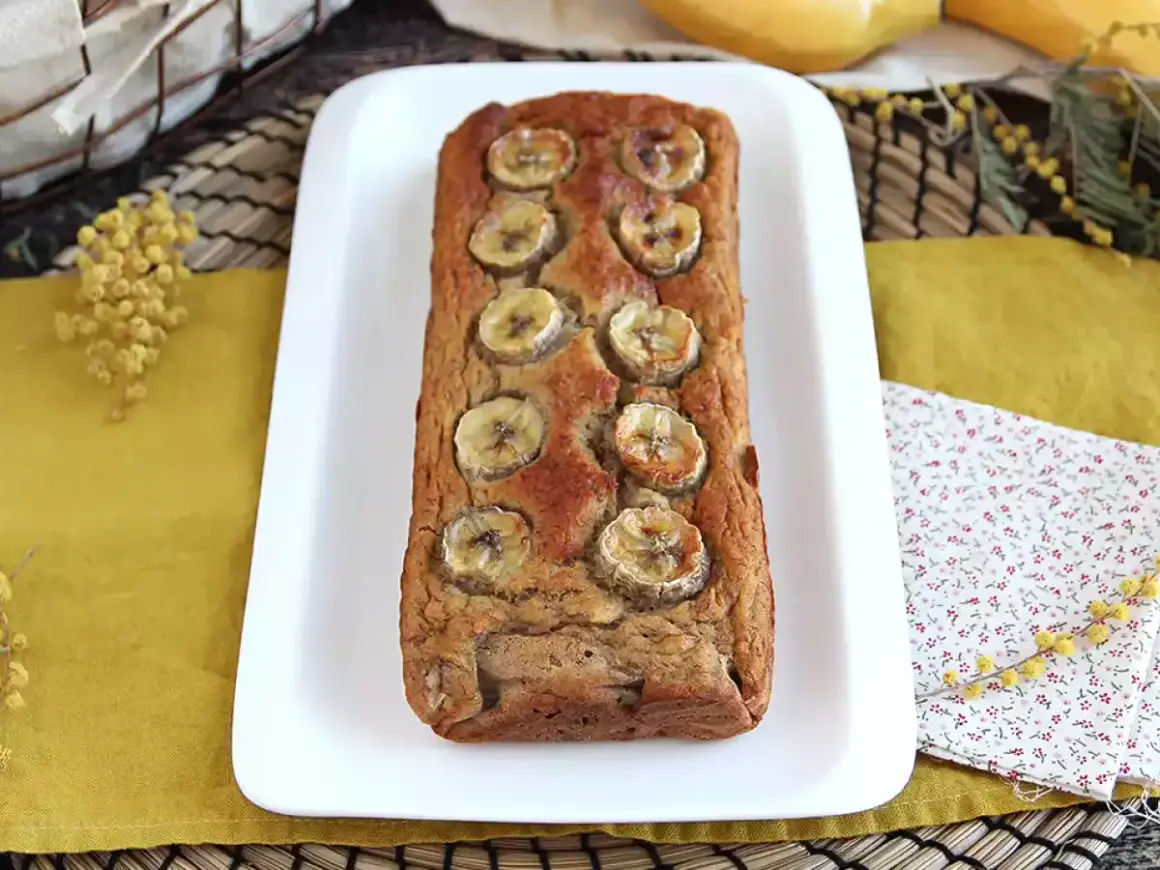 Delizioso banana bread light, decorato con fette di banana, su un piatto bianco, con fiori di mimosa sullo sfondo.