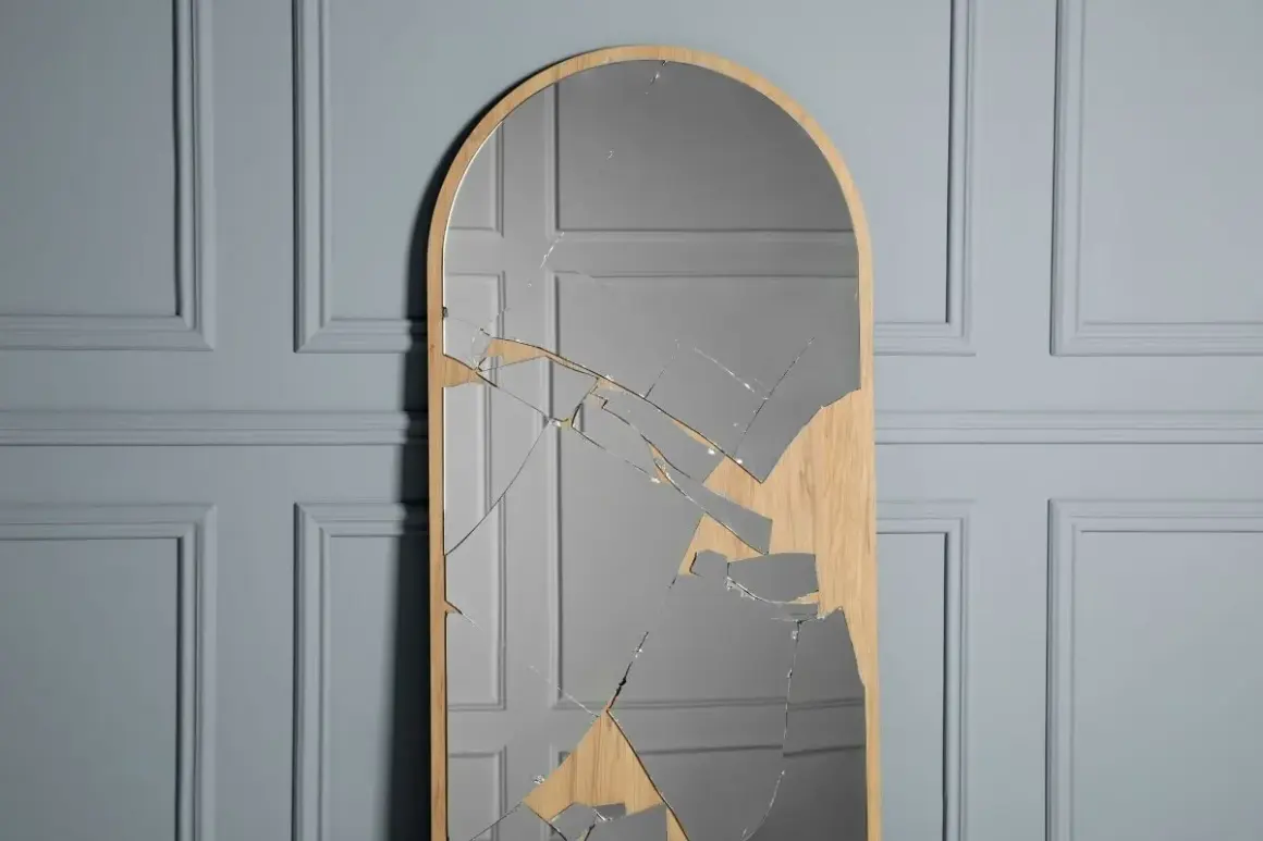 A tall, arched mirror with a wooden frame is shattered, with many glass shards reflecting a grey, paneled wall.