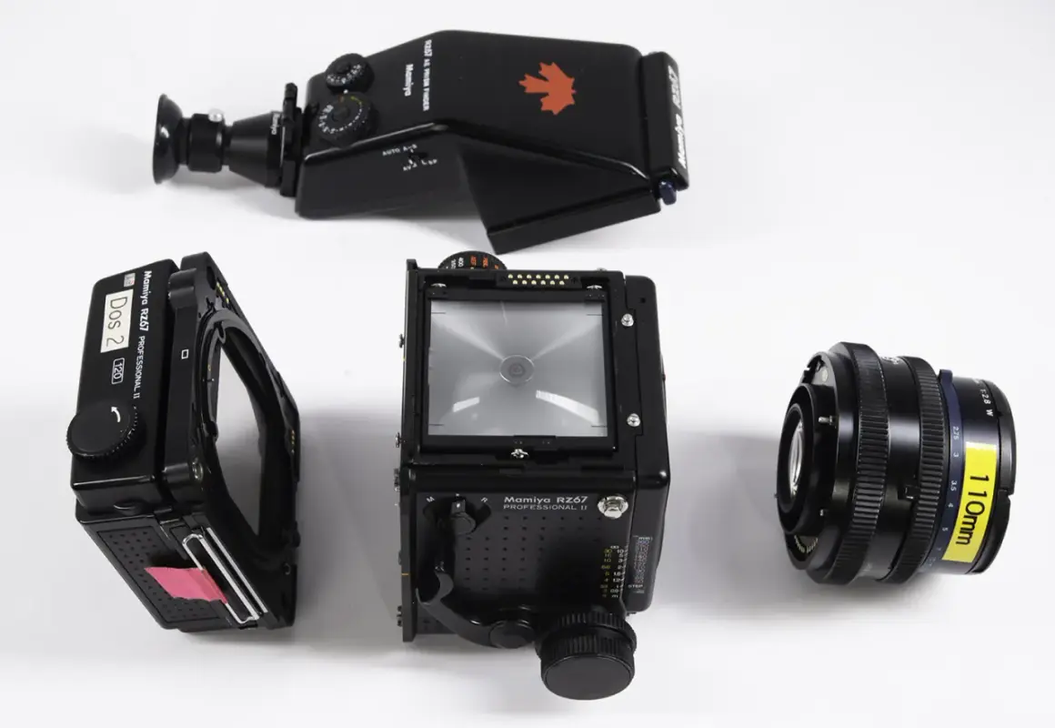 A Mamiya RZ67 camera body, film back, prism finder, and 110mm lens are laid out on a white surface.