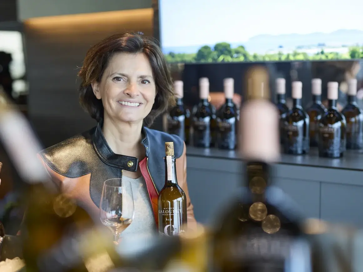 Frau mit Weinflasche und Glas, im Hintergrund Flaschenreihe und Landschaft.