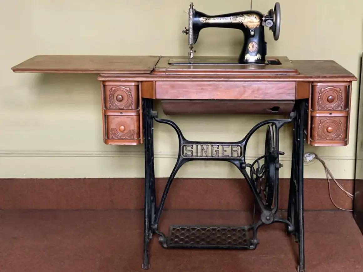 Antique Singer sewing machine on a wooden table with drawers and a treadle base.