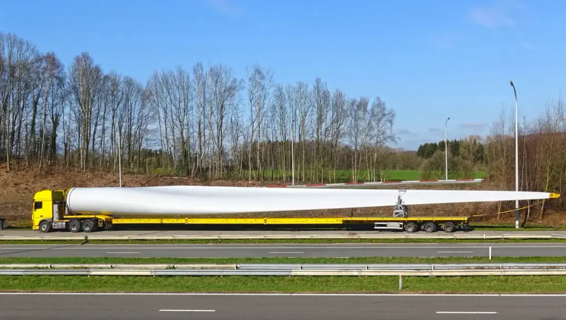 Żółta ciężarówka transportuje ogromne łopaty wiatraka. Ten pojazd nienormatywny pokonuje drogę, by dostarczyć element do budowy farmy wiatrowej.