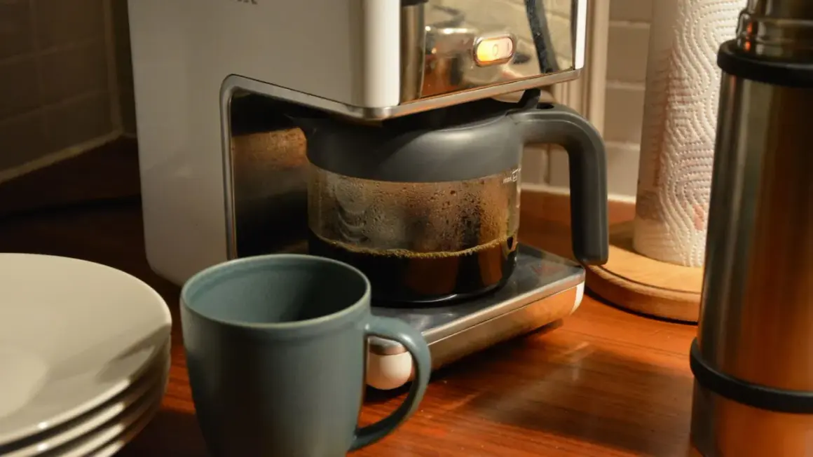 En kaffebryggare med en glaskanna full med färskt kaffe. Bredvid står en mugg och en stapel tallrikar. Man kan brygga presskaffe i kaffebryggare för en god start på dagen.
