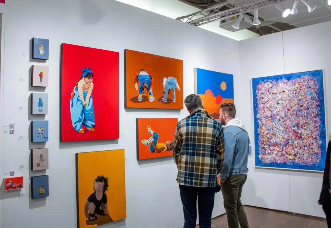 Two men examine colorful paintings at an art fair, perhaps contemplating their next acquisition as an art collector.