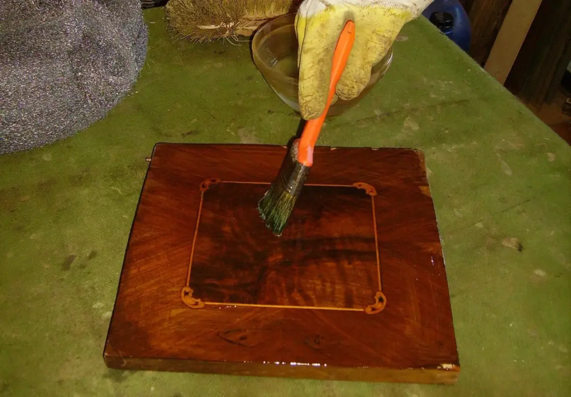Mano guantata applica liquido con pennello per sverniciare mobile legno. Accanto, paglietta metallica e spazzola.