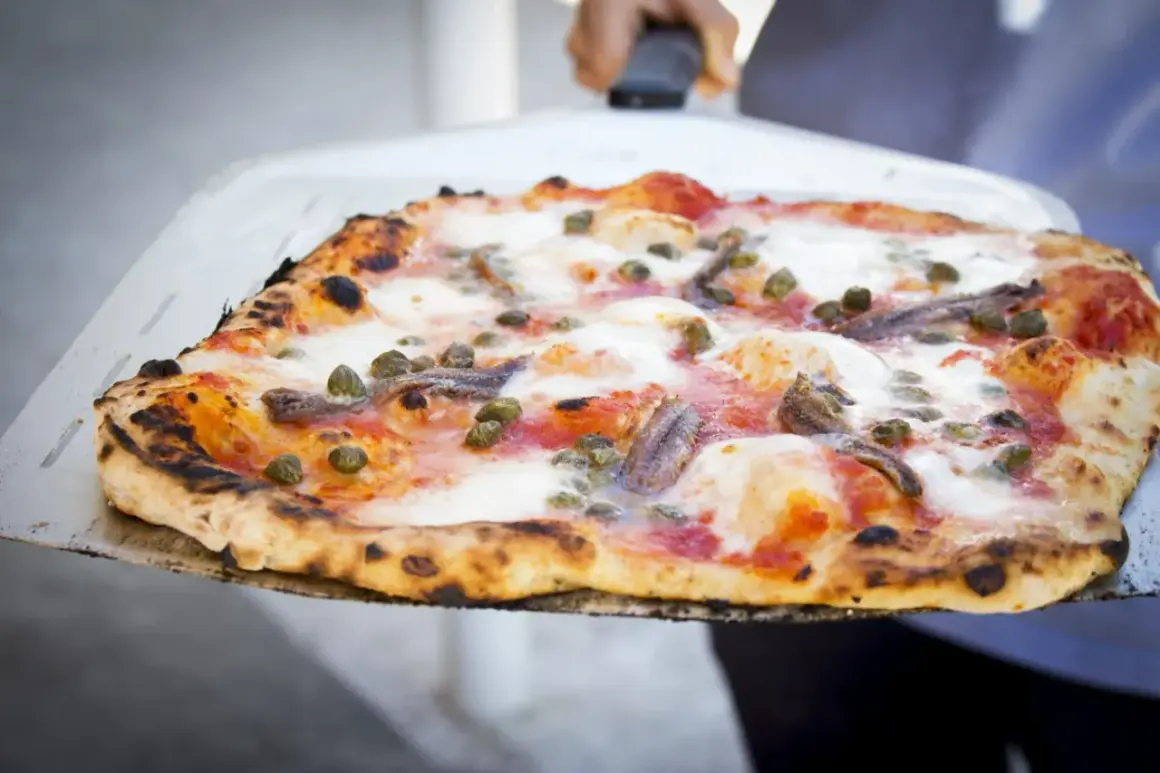 Pizza na redukcji z anchois i kaparami, wypiekana na cienkim cieście, prezentowana na łopacie.
