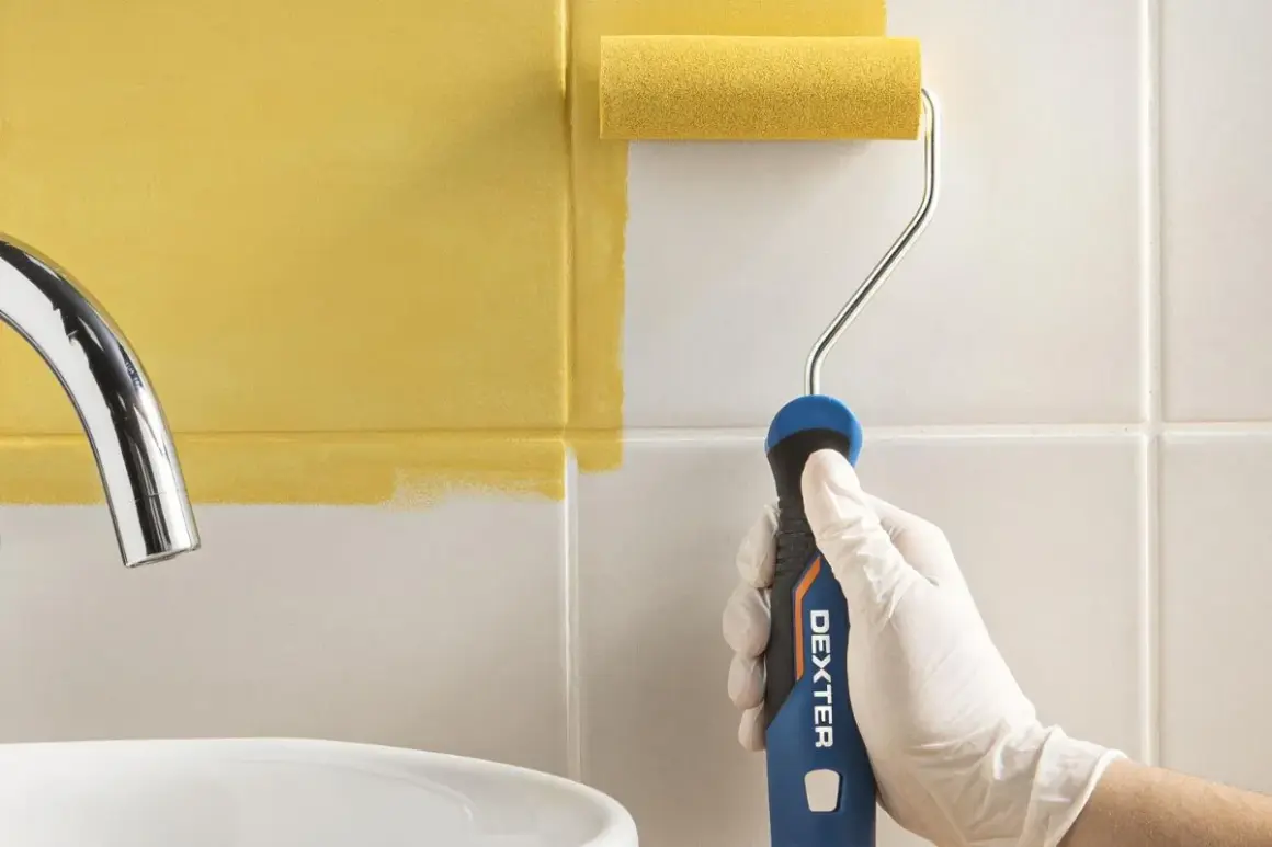 Mano con guante blanco pintando azulejos de baño de blanco a amarillo con rodillo.
